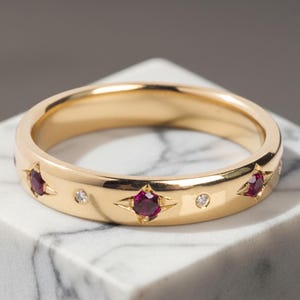 May include: A gold band ring featuring alternating round red rubies and small clear diamonds set into the metal. The gemstones are arranged in a repeating pattern around the circumference of the polished yellow gold band, creating a continuous design.