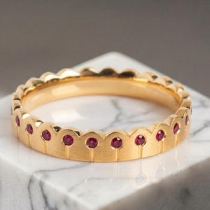 Puede incluir: Un anillo de oro con un diseño de borde festoneado a lo largo de la banda superior. El anillo está engastado con pequeños rubíes redondos de color rojo vibrante incrustados en cada festón, creando un patrón continuo de gemas en el contorno.
