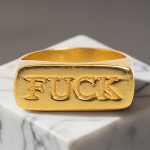 May include: A gold-toned metal ring featuring a rectangular face with the word FUCK embossed in capital letters across the front. The ring has a smooth, polished finish and a solid band design, showcasing a bold and prominent typographic detail.