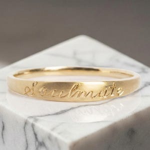 Könnte beinhalten: Ein dünner Goldring mit einer glatten, polierten Oberfläche. Das Wort soulmate ist in einer kursiven, fließenden Schriftart auf der Außenseite des Metallrings eingraviert, was diesem Schmuckstück ein zartes und raffiniertes Aussehen verleiht.