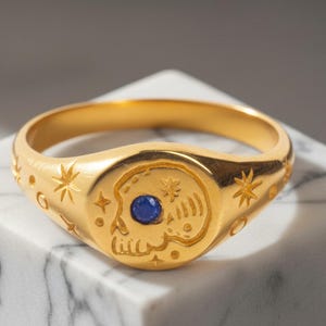 May include: Gold-colored signet ring featuring a skull design with a blue gemstone eye. The ring is adorned with star and crescent moon engravings. The ring is displayed on a white marble surface.