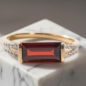 Puede incluir: Un anillo de oro con una piedra granate de color rojo intenso en talla baguette rectangular montada horizontalmente. La banda está acentuada con pequeñas piedras claras engastadas en pavé a lo largo de los hombros, creando un diseño dividido.