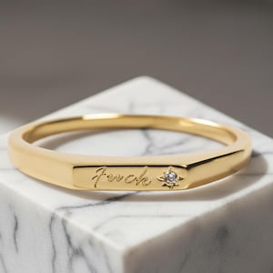 Puede incluir: Un anillo de banda en tono dorado con una superficie frontal plana y rectangular grabada con la palabra "fuck" en cursiva y un pequeño acento en forma de estrella con una piedra redonda transparente a la derecha del texto.
