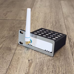 May include: A black electronic device with a silver front panel featuring a vertical white antenna. The device has a rectangular black housing with a grid of circular ventilation holes on top and two mounting slots on the left side panel.