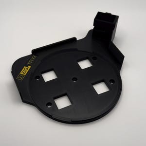 May include: A black, flat, circular plastic component featuring four square cutouts, several small circular mounting holes, and an extended rectangular housing at the top. The text 23 EIG, TFIVE is printed in yellow on the left side of the base.
