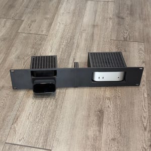 May include: A black rack-mount bracket featuring two rectangular cutouts, each topped with a grid-patterned ventilation section. One cutout holds a small black device, while the other contains a silver-toned electronic component with two visible ports.