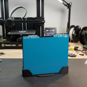 May include: A rectangular, bright blue 3D printer build plate held upright in a black plastic stand. The plate features white printed text and a snowflake icon along the top edge, with a smooth, solid-colored surface facing forward.