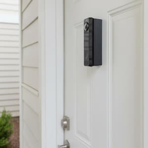 May include: A rectangular black video doorbell mounted on a white paneled door. The device features a circular camera lens at the top, a smooth dark surface, and a speaker grille at the bottom. The door includes a silver handle and deadbolt lock.