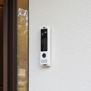 May include: A white rectangular smart video doorbell mounted on a textured wall. The device features a circular camera lens at the top, a vertical black display screen in the center, a circular button below the screen, and a speaker grille at the base.