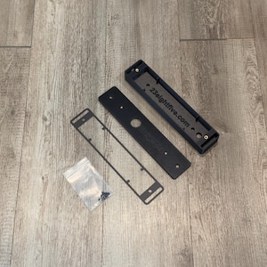 May include: A black rectangular mounting bracket kit consisting of a hollow base with the text 23eightfive.com, a flat cover plate, a thin frame, and a small clear plastic bag containing several metal screws for assembly and installation.