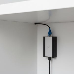 May include: A rectangular power injector with a silver center and black sides, featuring a small U-shaped logo. It is mounted vertically on a white wall, connected to a blue ethernet cable at the top and a black power cable at the bottom.