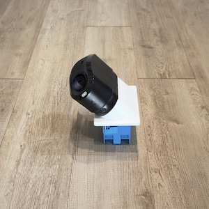 May include: A security camera featuring a black, rounded housing with a visible lens and a small white light, mounted onto a white rectangular base plate that connects to a blue, box-shaped electrical component with visible wiring ports.
