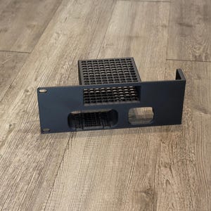 May include: A matte black metal rack mount panel featuring a rectangular perforated ventilation section on the top shelf and two distinct cutouts on the front face, including one square opening with a grid pattern and one smaller rectangular opening.