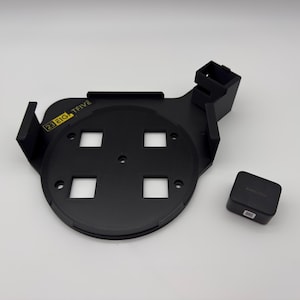 May include: A black circular mounting bracket with four square cutouts, raised side supports, and a small rectangular attachment point. The text 23 EIG TFIVE is printed in yellow on the top surface. A separate small black square block is included.