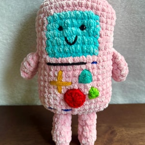 May include: A handmade pink crochet plush toy in the shape of a retro video game console. The toy features a teal screen, black eyes and mouth, and colorful buttons. The arms and legs are short and stubby. The toy is made of soft, textured yarn.