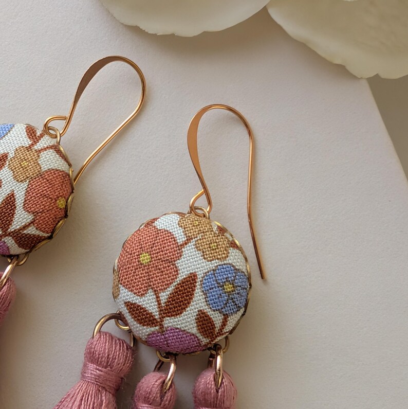 May include: Close-up of floral fabric earrings with gold-colored hooks and pink tassels. The earrings feature a round, fabric-covered design with a pattern of flowers in shades of orange, blue, and purple. The tassels are a soft pink color.