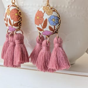 May include: A pair of dangle earrings featuring floral fabric pendants with gold-tone metal trim. Each earring has three dusty rose tassels suspended below. The fabric has orange, blue, and purple flowers.