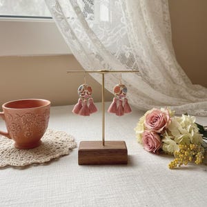 May include: A pair of floral earrings with pink tassels displayed on a gold stand with a wooden base. The earrings have a circular design with a colorful floral pattern. A peach-colored teacup and a bouquet of roses are in the background.