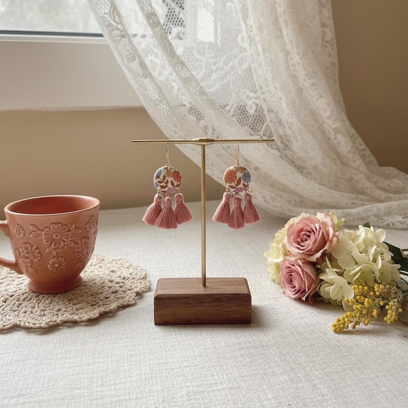 May include: A pair of floral earrings with pink tassels displayed on a gold stand with a wooden base. The earrings have a circular design with a colorful floral pattern. A peach-colored teacup and a bouquet of roses are in the background.