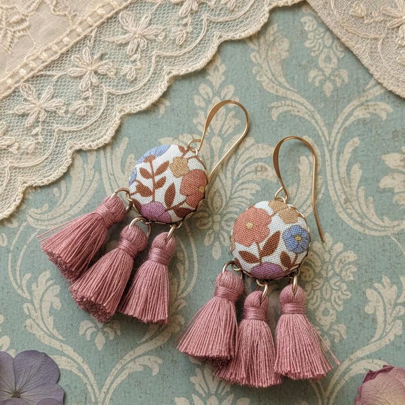 May include: A pair of floral fabric earrings with gold-colored hooks and pink tassels. The earrings feature a round, fabric-covered button with a floral pattern in shades of pink, orange, and blue. Each earring has three pink tassels hanging below.