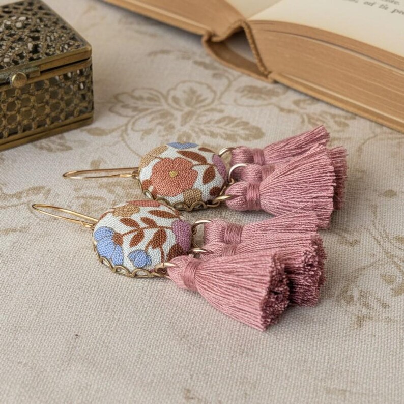 May include: A pair of gold-toned earrings with floral fabric-covered button tops and pink tassel drops. The fabric features a pattern of blue, orange, and brown flowers. The earrings are displayed on a patterned surface.