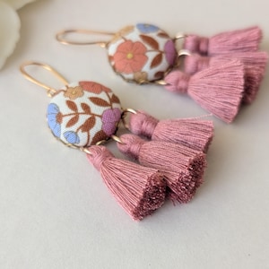 May include: A pair of dangle earrings with floral fabric-covered button tops and pink tassel accents. The earrings have gold-colored ear wires. The fabric features a pattern of flowers in shades of orange, blue, and purple on a white background.