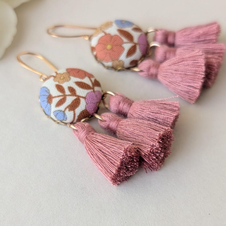 May include: A pair of dangle earrings with floral fabric-covered button tops and pink tassel accents. The earrings have gold-colored ear wires. The fabric features a pattern of flowers in shades of orange, blue, and purple on a white background.