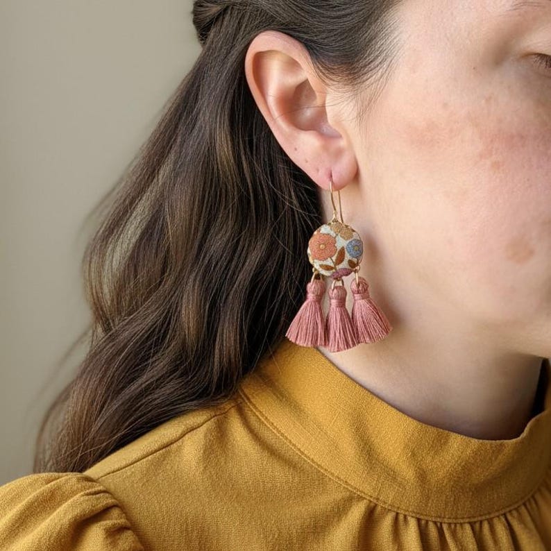May include: Floral embroidered earrings with pink tassels. The earrings feature a round, fabric-covered design with a colorful floral pattern in shades of pink, blue, and orange. The tassels are a dusty rose color.
