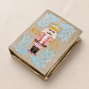 Peut inclure: Une couverture de livre en velours taupe ornée d'un casse-noisette brodé portant une couronne dorée, entouré de branches feuillues bleu clair. Le mot NOEL est cousu en fil doré, le casse-noisette étant placé entre les lettres O et E.