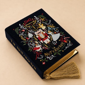 May include: A dark blue velvet book titled The Nutcracker with gold foil lettering on the spine. The cover features colorful embroidery of a nutcracker, a mouse, and a dancer surrounded by festive greenery, candy canes, and ribbons with a gold tassel.