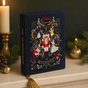 Puede incluir: Un libro azul marino con un diseño bordado que presenta un cascanueces, un ángel y otros elementos navideños. El título "The Nutcracker" se muestra en el lomo y la portada. Una borla dorada cuelga del libro.