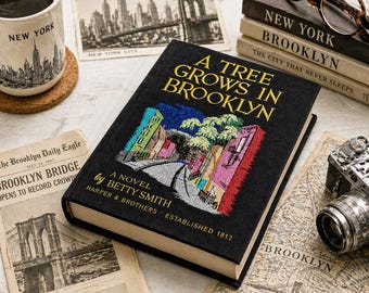 May include: A black hardcover book titled "A Tree Grows in Brooklyn" with colorful embroidered artwork. The image includes a coffee mug, vintage newspaper clippings, and a small camera, evoking a literary theme.