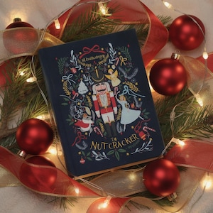 Puede incluir: Un libro azul marino titulado "The Nutcracker" con una colorida ilustración del Cascanueces, una bailarina y otros elementos festivos. El libro está rodeado de cintas rojas y doradas, adornos rojos y luces navideñas.
