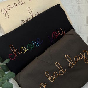 Könnte beinhalten: Drei gefaltete Sweatshirts in Creme, Schwarz und Dunkelgrau. Das cremefarbene Sweatshirt hat den gestickten Text "good vibes only" in Braun. Das schwarze Sweatshirt hat "choose you" in Regenbogenfarben gestickt. Das dunkelgraue Sweatshirt hat "no bad days" in Gold gestickt.