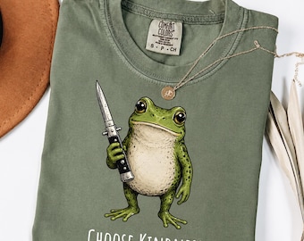 May include: An olive green t-shirt featuring a graphic of a green frog holding a silver switchblade knife. Below the frog, the text reads "CHOOSE KINDNESS" in white letters. The shirt has a Comfort Colors brand label inside the collar.