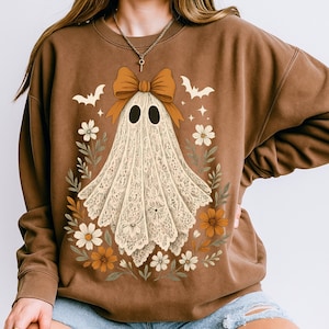 May include: Brown sweatshirt featuring a ghost design with a large orange bow, black eyes, and a floral wreath. The ghost is made of a lace-like material. Bats and stars are also included in the design. A fall-themed, Halloween-inspired design.