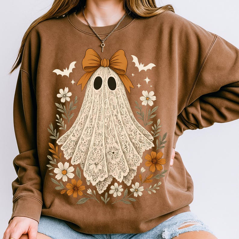 May include: Brown sweatshirt featuring a ghost design with a large orange bow, black eyes, and a floral wreath. The ghost is made of a lace-like material. Bats and stars are also included in the design. A fall-themed, Halloween-inspired design.