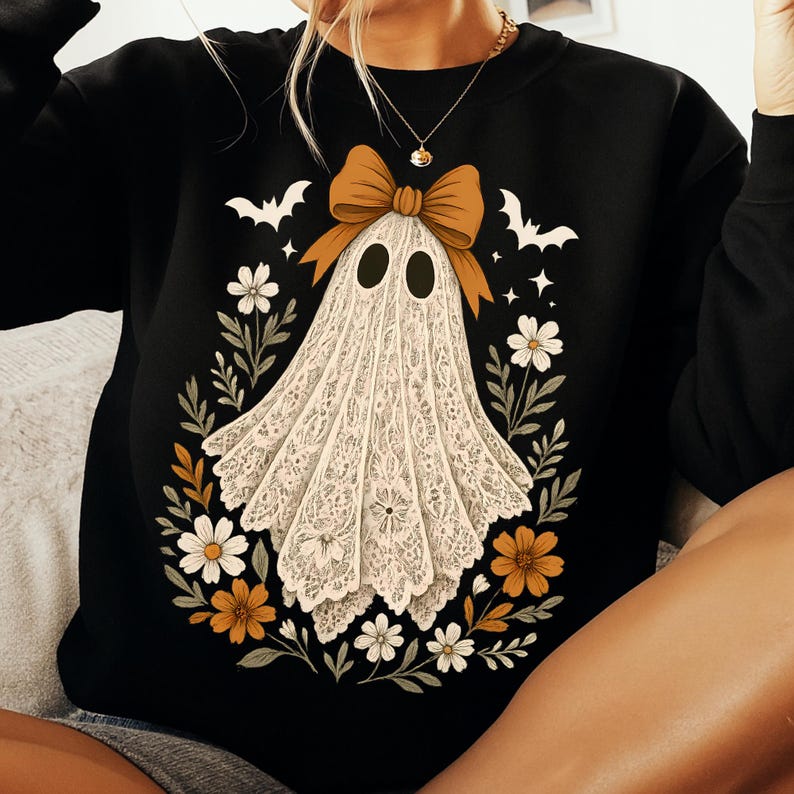 May include: Black sweatshirt featuring a white ghost design with a tan bow, surrounded by flowers and bats. The ghost has two black oval eyes and is draped in a lace-like fabric. The design has a Halloween theme.