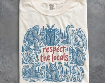 May include: A cream-colored t-shirt featuring a blue graphic print of various cryptids and the red text "respect the locals" in the center. The design includes illustrations of Bigfoot, a mothman, a sea serpent, an alien, and other creatures.