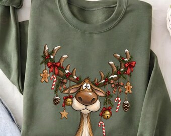 May include: An olive green crewneck sweatshirt featuring a graphic of a reindeer with large antlers decorated with pine branches, red bows, gingerbread men, candy canes, pinecones, gold stars, and jingle bells hanging from the antlers.