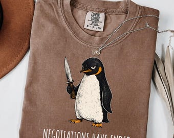 May include: A brown short-sleeve t-shirt featuring a graphic of a penguin holding a knife. The text "NEGOTIATIONS HAVE ENDED" is printed in white below the penguin. The shirt has a Comfort Colors label at the neck and a crew neckline.