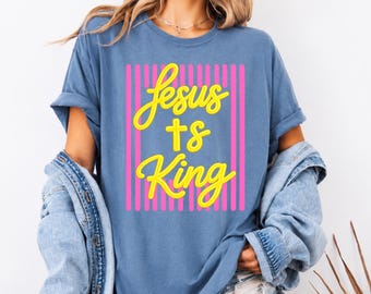 May include: A blue short-sleeved t-shirt featuring a graphic design with vertical pink stripes and the yellow cursive text "Jesus is King" centered on the front. The shirt has a crew neckline and a relaxed fit, worn with a denim jacket over the shoulders.