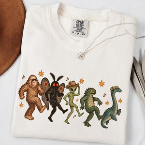 May include: An off-white crew neck t-shirt with a Comfort Colors label. The graphic features a line of five mythical creatures including Bigfoot, Mothman, an alien in a cowboy hat, a swamp monster, and a sea serpent, all surrounded by orange stars.