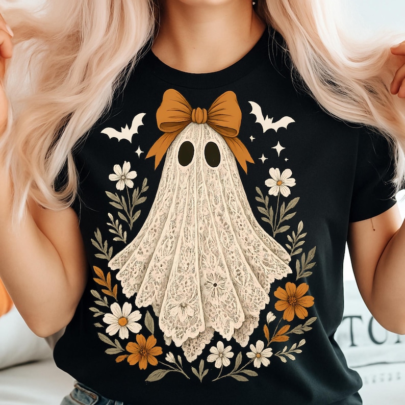May include: Black t-shirt featuring a ghost design. The ghost is adorned with a bow and surrounded by flowers and bats. The ghost is made of lace. The design includes white, orange, and brown colors, perfect for Halloween.