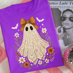 May include: A purple sweatshirt featuring a ghost design. The ghost has a lace-like appearance, a brown bow, and is surrounded by flowers, bats, and stars. The design is centered on the shirt.