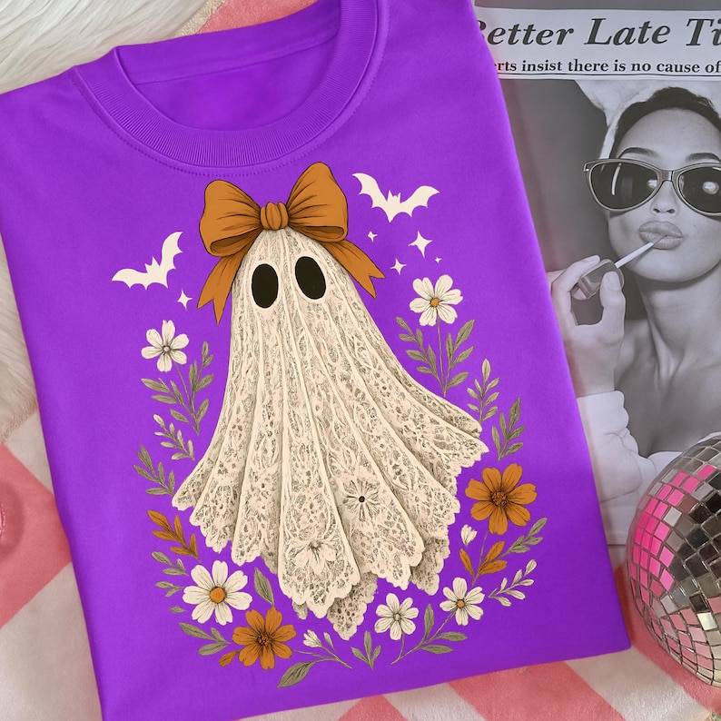 May include: A purple sweatshirt featuring a ghost design. The ghost has a lace-like appearance, a brown bow, and is surrounded by flowers, bats, and stars. The design is centered on the shirt.