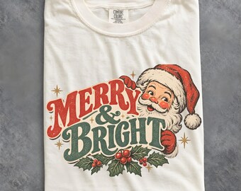 May include: An off-white crew neck t-shirt featuring a graphic of Santa Claus with the text MERRY & BRIGHT in red and green lettering. The design includes holly leaves and berries, along with small star accents, printed on the front of the shirt.