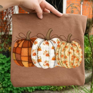May include: A brown tote bag featuring three decorative pumpkins. The pumpkins have different patterns: patchwork, white with orange pumpkins, and floral. Each pumpkin has a brown stem and is connected by a brown vine.
