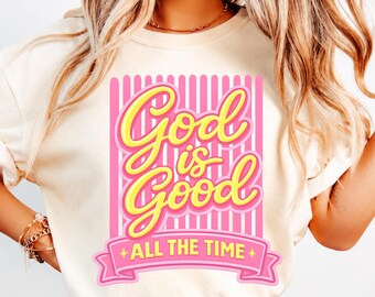 May include: A cream-colored short-sleeve t-shirt featuring a graphic print with vertical pink stripes. The text "God is Good" appears in large yellow script, with "ALL THE TIME" printed in yellow on a pink banner at the bottom of the design.
