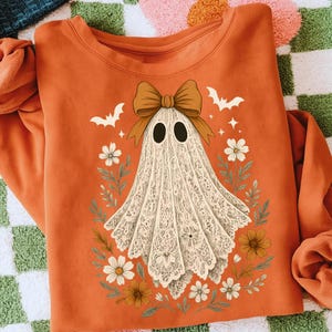 May include: Orange sweatshirt featuring a ghost design. The ghost is depicted with a white lace-like body, black oval eyes, and a brown bow. The design includes bats, flowers, and leaves, creating a Halloween theme.